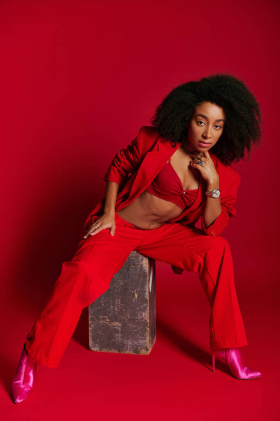 appealing sexy african american woman in red attire sitting and looking at camera, fashion concept - 写真・画像