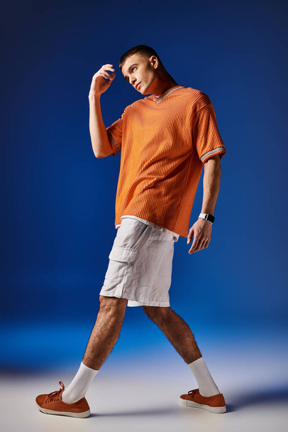 Full length image of handsome man in orange shirt and white shorts posing on blue backdrop - Foto, afbeelding