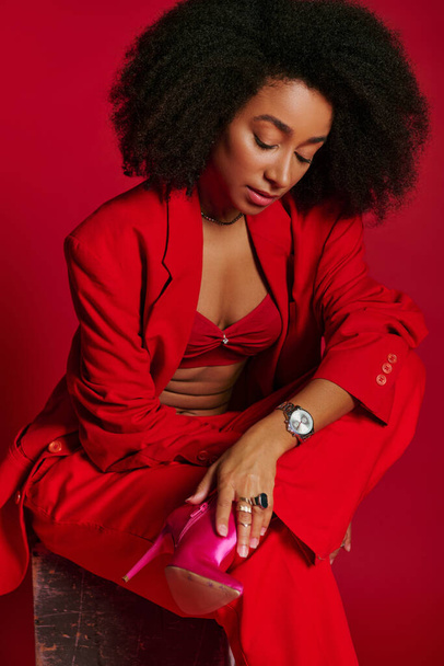 sexy donna afroamericana in abito rosso seduta e guardando altrove su sfondo rosso, concetto di moda - Foto, immagini
