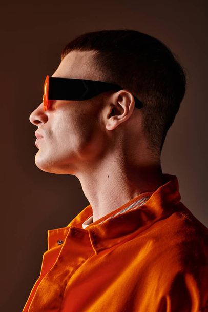 Πλάγια εικόνα του κομψού άνδρα σε πορτοκαλί πουκάμισο και γυαλιά ηλίου σε καφέ φόντο - Φωτογραφία, εικόνα
