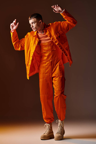Fashion full length shot van stijlvolle man in oranje jumpsuit en jas poseren op bruine achtergrond - Foto, afbeelding