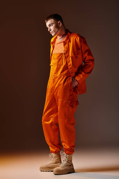 Full length portret van modieuze man in oranje jumpsuit en jasje staand op bruine achtergrond - Foto, afbeelding