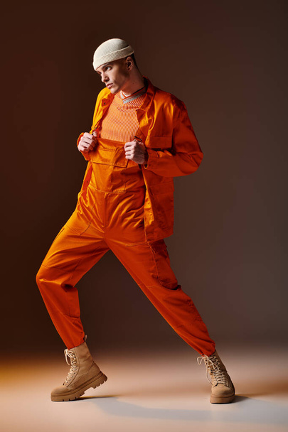 Fashion shot van stijlvolle man in oranje jumpsuit en jas, beige beanie op bruine achtergrond - Foto, afbeelding