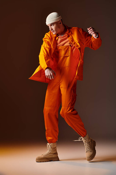 Volledige shot van de mens in oranje jumpsuit en jas, beige beanie poseren op bruine achtergrond - Foto, afbeelding