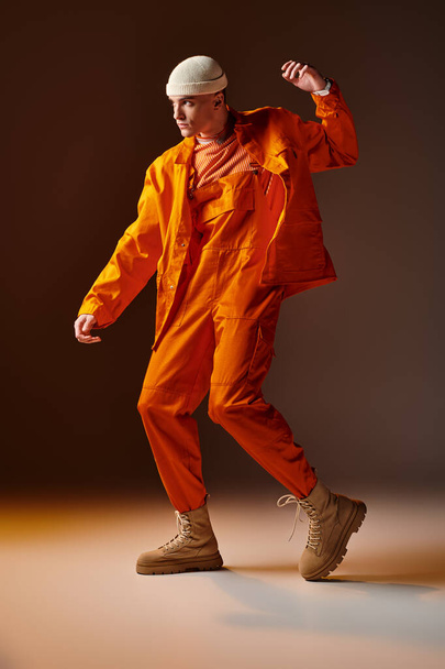 Молодой красивый мужчина в оранжевом комбинезоне и куртке, бежевая шапочка позирует на коричневом фоне - Фото, изображение