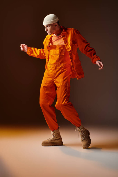 Stylový muž v oranžové kombinéze a bundě, béžová beanie pózuje na hnědém pozadí - Fotografie, Obrázek