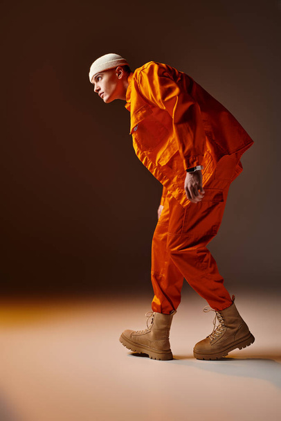 Side view of handsome man in orange jumpsuit and jacket, beige beanie standing on brown background - Фото, изображение