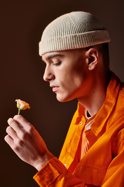 Πλευρική άποψη του άντρα της μόδας σε πορτοκαλί στολή φορώντας μπεζ σκούφο κρατώντας λουλούδι στο χέρι - Φωτογραφία, εικόνα