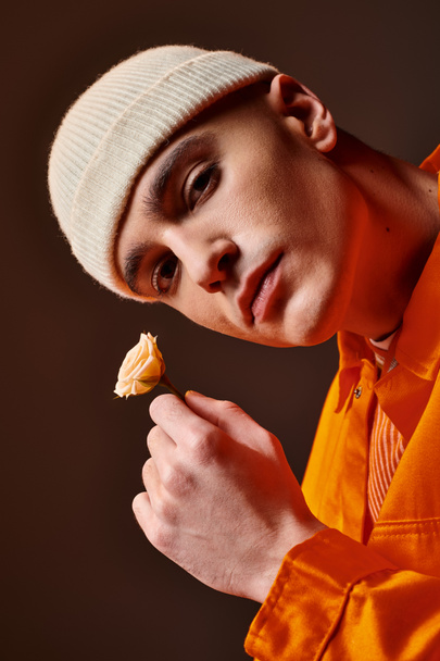 bel homme en tenue orange portant bonnet beige tenant fleur dans la main et regardant la caméra - Photo, image