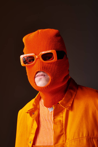 Elegante chico en traje naranja con mascarilla de pasamontañas y gafas de sol naranjas sobre fondo marrón - Foto, imagen