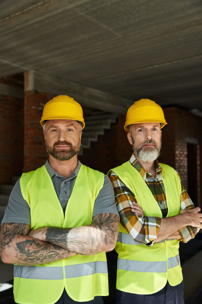 dos hombres barbudos de buen aspecto en chalecos de seguridad y cascos mirando a la cámara, constructores de cabañas - Foto, Imagen