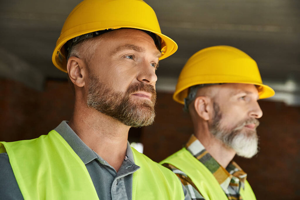 homens barbudos atraentes em capacetes de segurança e coletes posando e olhando para longe, construtores de casas de campo - Foto, Imagem