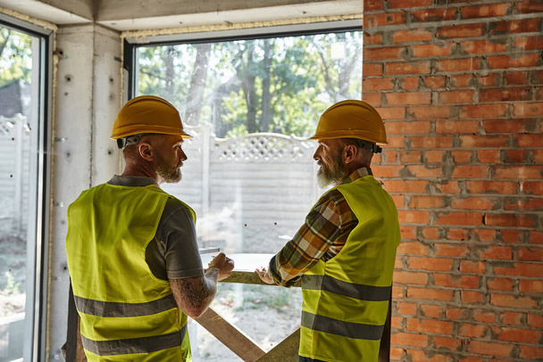 two hardworking bearded men in safety vests and helmets on construction site, cottage builders - Photo, Image