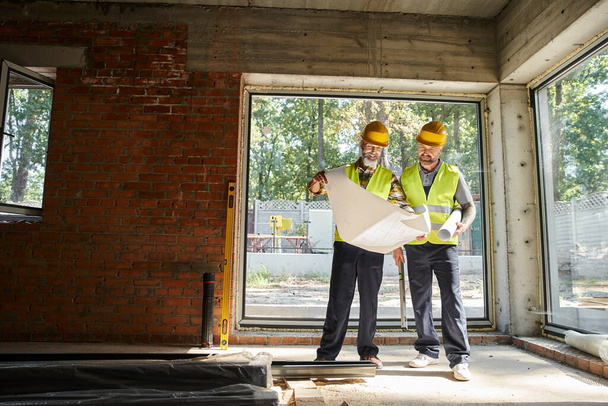 hommes barbus joyeux dans des casques de sécurité regardant le plan tandis que sur le chantier de construction, constructeurs - Photo, image