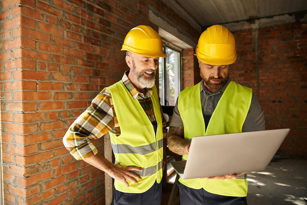veselí oddaní stavební dělníci v bezpečnostních vestách pracující s notebookem na místě, stavitelé chalup - Fotografie, Obrázek