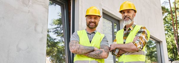 jolly workers in safety vests with arms crossed looking at camera, cottage builders, banner - Photo, Image