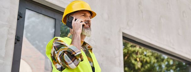 radosny dedykowany człowiek w kasku bezpieczeństwa rozmawiający przez telefon na placu budowy, budowniczy domków, baner - Zdjęcie, obraz