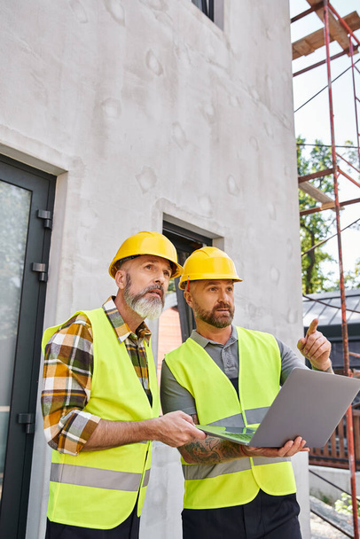 handsome bearded men in safety vests and helmets working with laptop on construction site, builders - Photo, Image