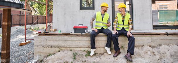 радісні будівельники котеджів у безпечних шоломах сидять на ганку і дивляться один на одного, банер - Фото, зображення