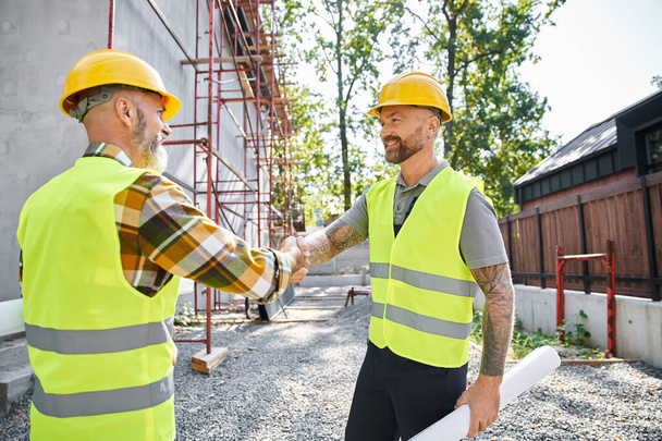 Neşeli inşaat işçileri el sıkışıyor ve birbirlerine gülümsüyorlar, kulübe inşaatçıları. - Fotoğraf, Görsel