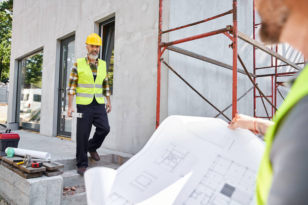 focus on bearded jolly cottage builder with level looking at his blurred colleague with blueprint - Photo, Image