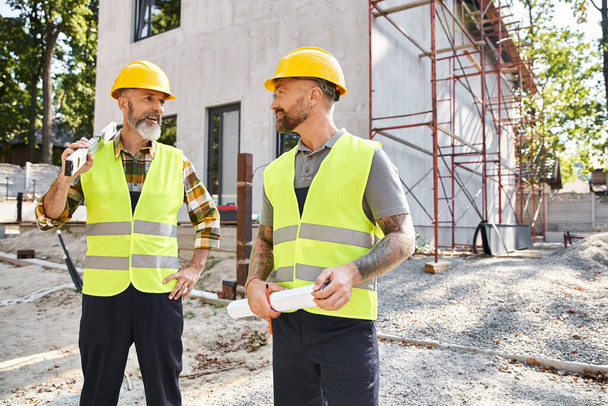 atractivos trabajadores de la construcción alegres con nivel y plano mirándose entre sí, constructores - Foto, Imagen