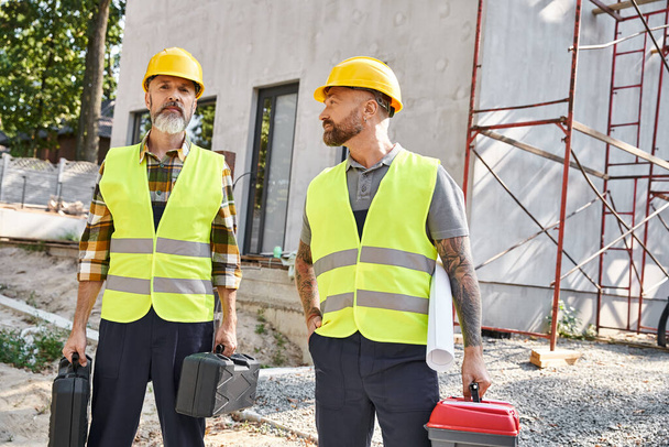 dwóch atrakcyjnych mężczyzn w kaskach ochronnych i kamizelkach pozujących ze skrzynkami z narzędziami i planami, budowniczowie domków - Zdjęcie, obraz