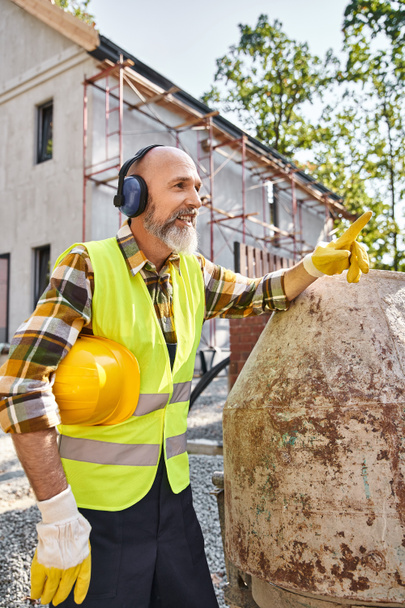 веселий будівельник котеджу в рукавичках безпеки і жилеті позує з навушниками і шоломом на місці - Фото, зображення