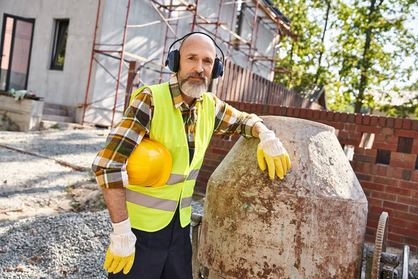 cheerful cottage builder in safety gloves and vest with headphones and helmet smiling at camera - Photo, Image