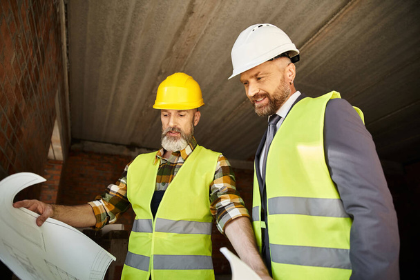 cheerful cottage builder and businessman in safety vests discussing blueprint on construction site - Photo, Image