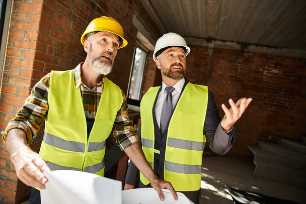веселый строитель коттеджей и бизнесмен с чертежом обсуждают свою работу на строительной площадке - Фото, изображение