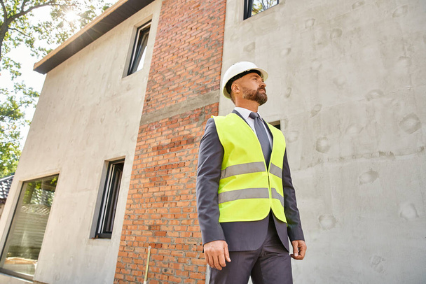 Engagierter Geschäftsmann in Warnweste und Helm posiert auf Baustelle und schaut weg - Foto, Bild