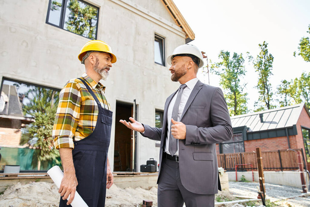 jolly cottage builder and architect in suit and overalls with blueprint discussing construction - Photo, Image