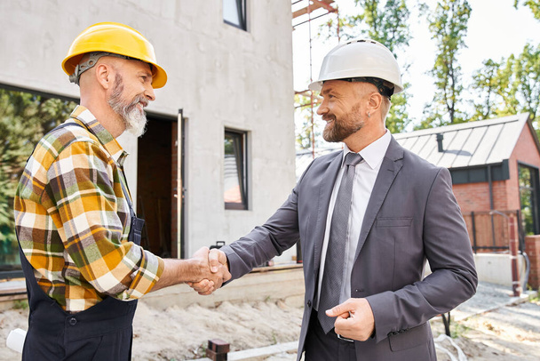 joyous handsome architect and builder in overalls and suit with safety helmets shaking hands happily - Photo, Image