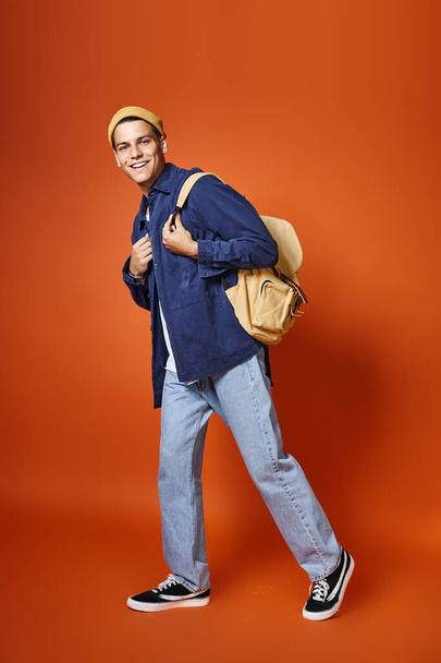 happy man in his 20s with grey eyes and backpack walking on terracotta background - Photo, Image