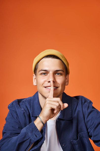 портрет улыбающегося мужчины в возрасте 20 лет, показывающий знак молчания на фоне терракотовых - Фото, изображение