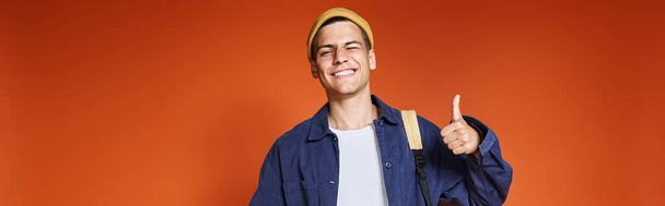 bannière de jeune homme heureux en chapeau debout avec sac à dos et montrant comme sur fond de terre cuite - Photo, image