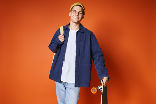 charismatische jongeman in casual outfit staande met skateboard tegen terracotta achtergrond - Foto, afbeelding