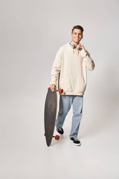 charmante man in witte hoodie en koptelefoon lopen met skateboard op lichte achtergrond - Foto, afbeelding
