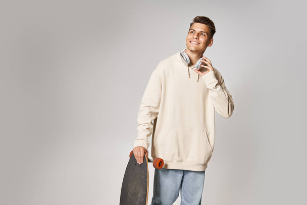усміхнений молодий чоловік в навушниках зі скейтбордом дивиться вгору на світлому фоні - Фото, зображення