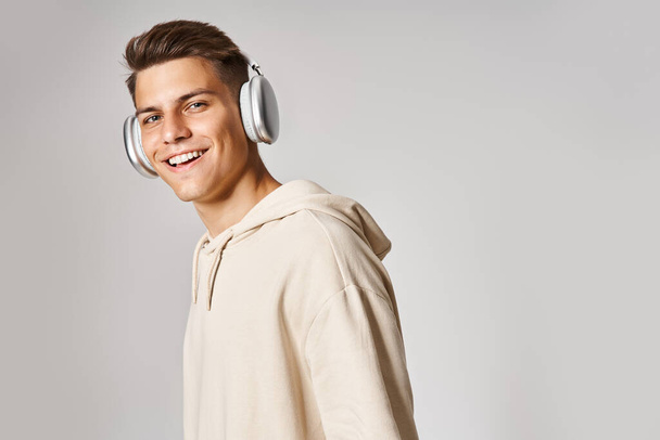 jovem atraente com cabelo castanho em fones de ouvido sorrindo para câmera contra fundo leve - Foto, Imagem