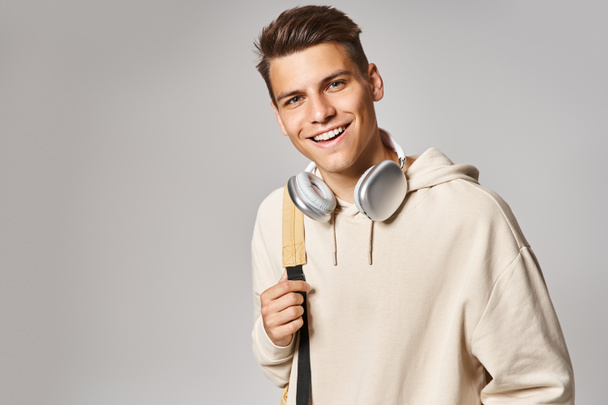 atractivo joven estudiante en auriculares y atuendo casual con mochila sobre fondo gris - Foto, Imagen