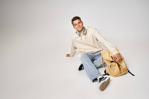 красивий молодий чоловік в навушниках і повсякденному вбранні сидить з рюкзаком на сірому фоні - Фото, зображення