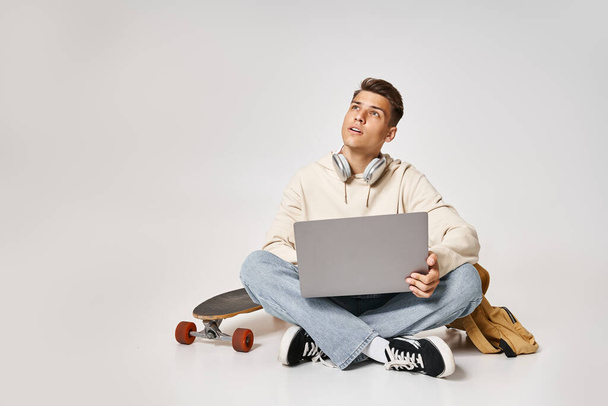 όμορφος μαθητής σε ακουστικά δικτύωσης σε φορητό υπολογιστή και κάθεται με σακίδιο πλάτης και skateboard - Φωτογραφία, εικόνα