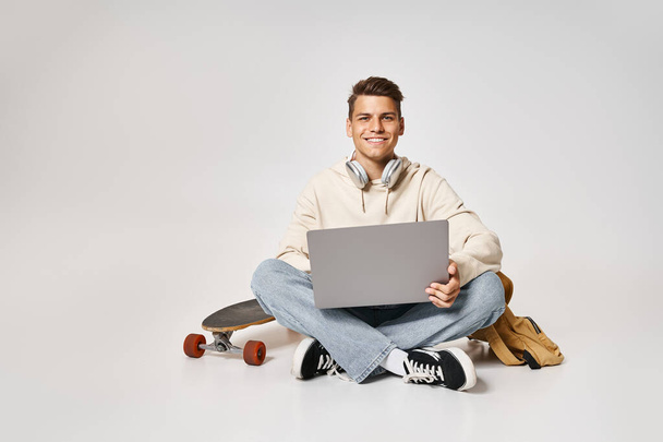 улыбающийся молодой студент в наушниках, подключенных к ноутбуку и сидящий с рюкзаком и скейтбордом - Фото, изображение