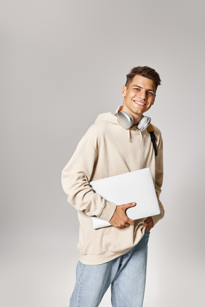 20 'li yaşlarda, laptoplu ve kulaklıklı yakışıklı bir öğrenci gri arka planda yürüyor. - Fotoğraf, Görsel