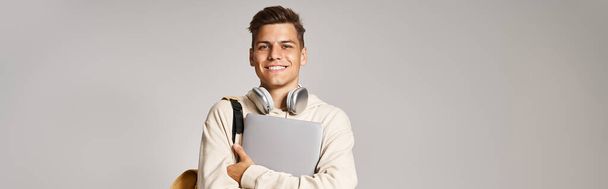 οριζόντια φωτογραφία του μαθητή στα 20 με φορητό υπολογιστή και ακουστικά με αυτοπεποίθηση στέκεται σε γκρι φόντο - Φωτογραφία, εικόνα