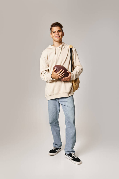 fröhliche Studentin im lässigen Outfit mit Rucksack mit Rugby-Ball auf grauem Hintergrund - Foto, Bild