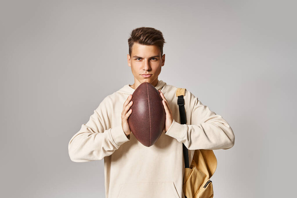 homme confiant dans sa vingtaine avec sac à dos tenant la balle de rugby avec les mains sur fond gris - Photo, image