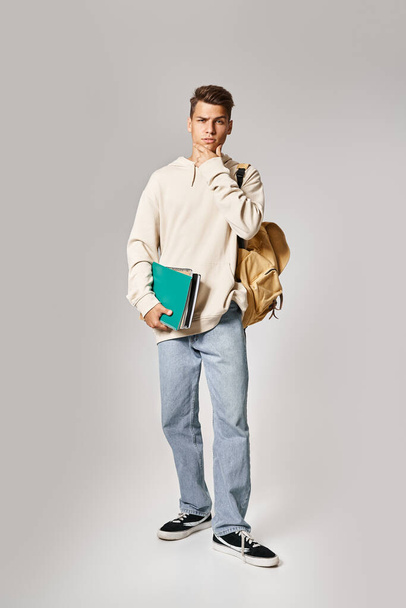 carismático joven estudiante con mochila sosteniendo notas y tocando la mandíbula con las manos - Foto, Imagen